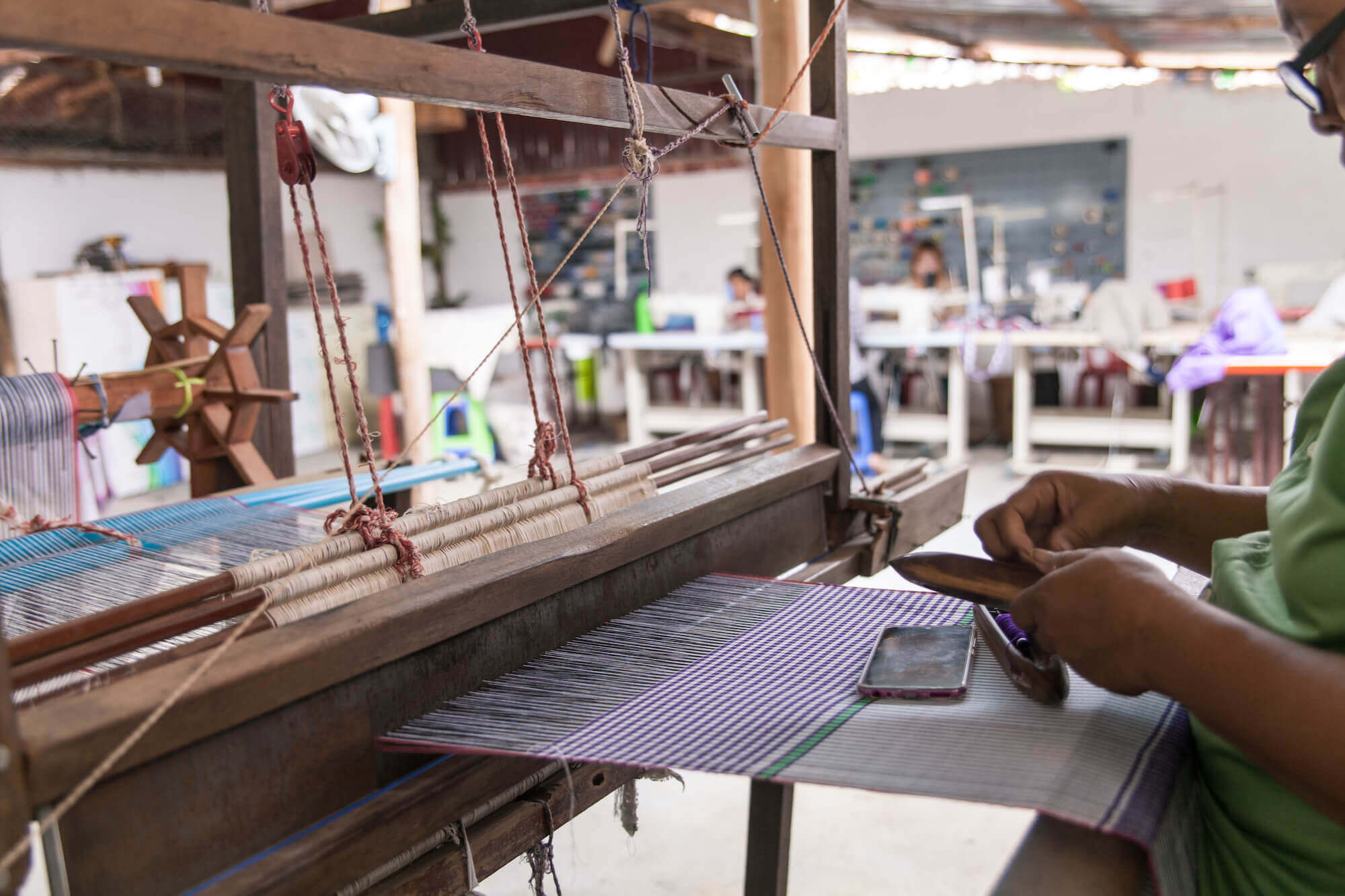 Photo of one of the wonderful local women in the production centre in Cambodia weaving on a large wooden weaving machine while watching television on her phone