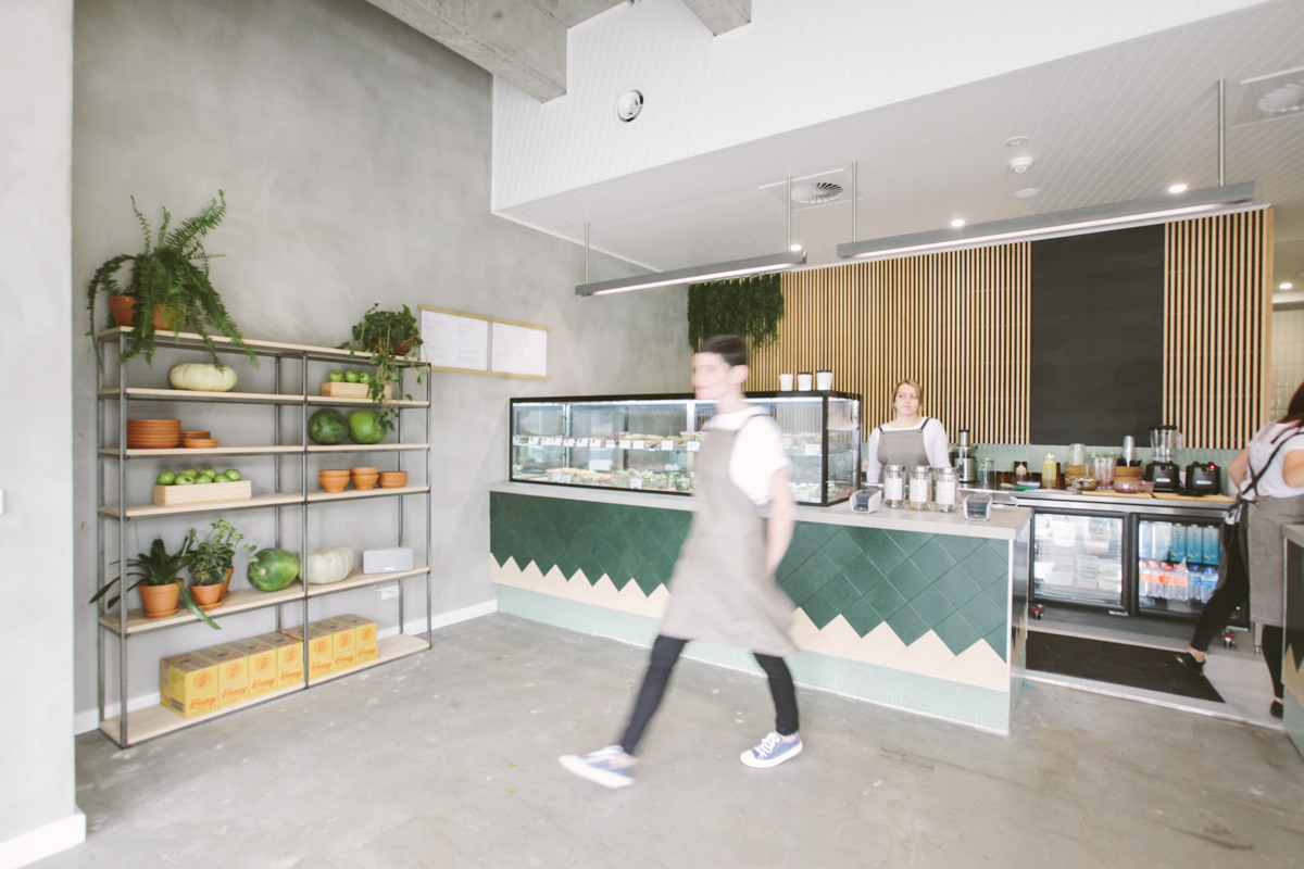 Photo of one of the client projects, this one of a cafe with polished cement floors showing a staff member blurred walking through the photo and in the background a green tiled counter with wood panelling on the wall