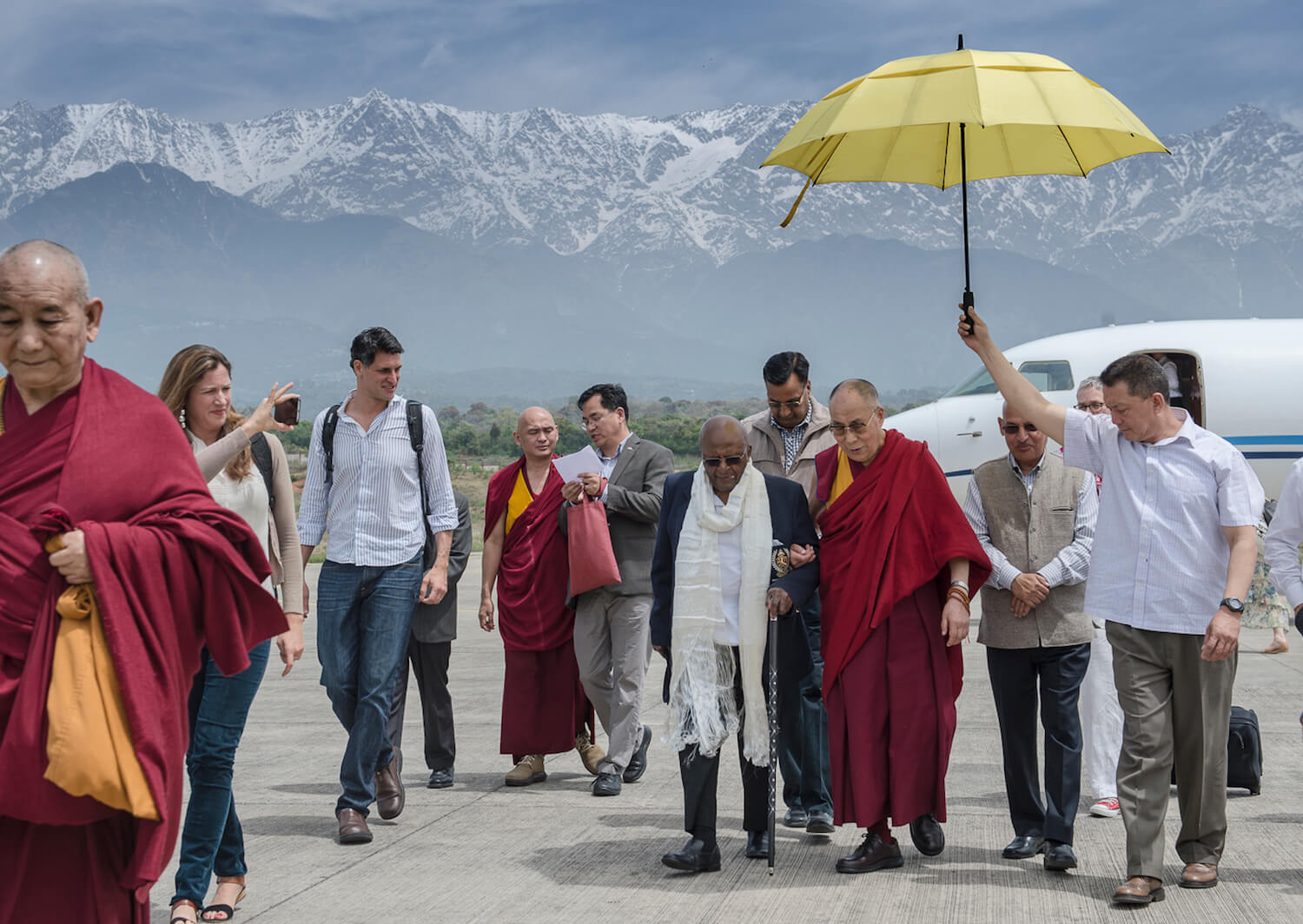 A photo from the trip to India with monks and the writing team. Desmond Tutu and the Dalai Lama are walking together under a yellow umbrella while Doug Abrams is walking next to them. There are snow capped mountains in the background.