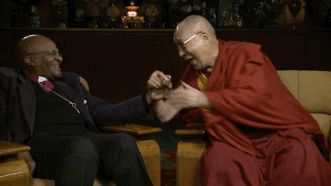 The Dalai Lama taking the hand of Desmond Tutu agaist his cheek in a warm gesture while laughing