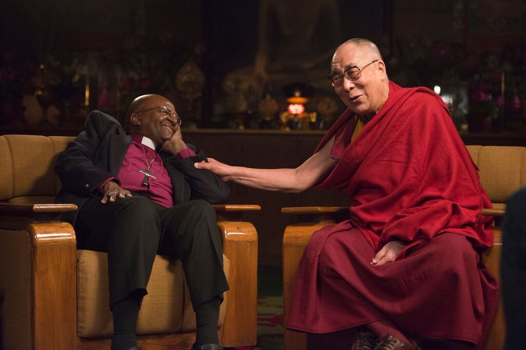 His Holiness the Dalai Lama and Archbishop Desmond Tutu are sitting together on two large, comfortable wooden chairs. Desmond Tutu is looking at the Dalai Lama while smiling. The Dalai Lama has his hand placed on the arm of Desmond Tutu in a gesture of friendship and is laughing with his eyes closed.