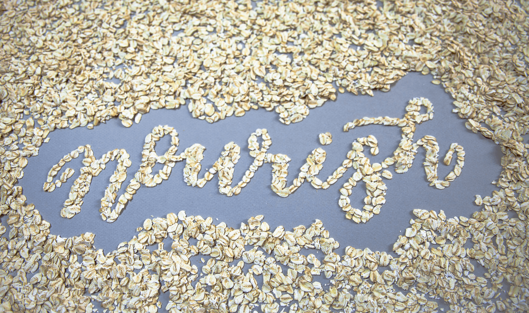 The word nourish is shown made from light colored oats in cursive form on a blue-grey background