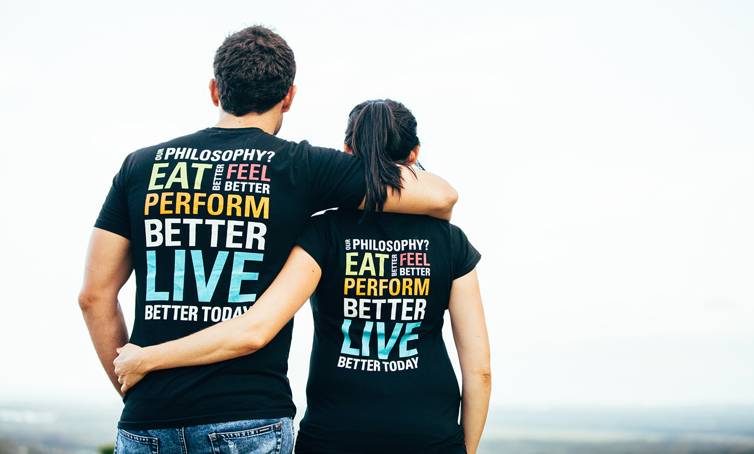 The two founders of Howzya stand with their backs to the camera, outdoors, wearing black shirt with bold sans serif typography in multiple colors that reads, Our philosphy? Eat better, feel better, perform better, live better today