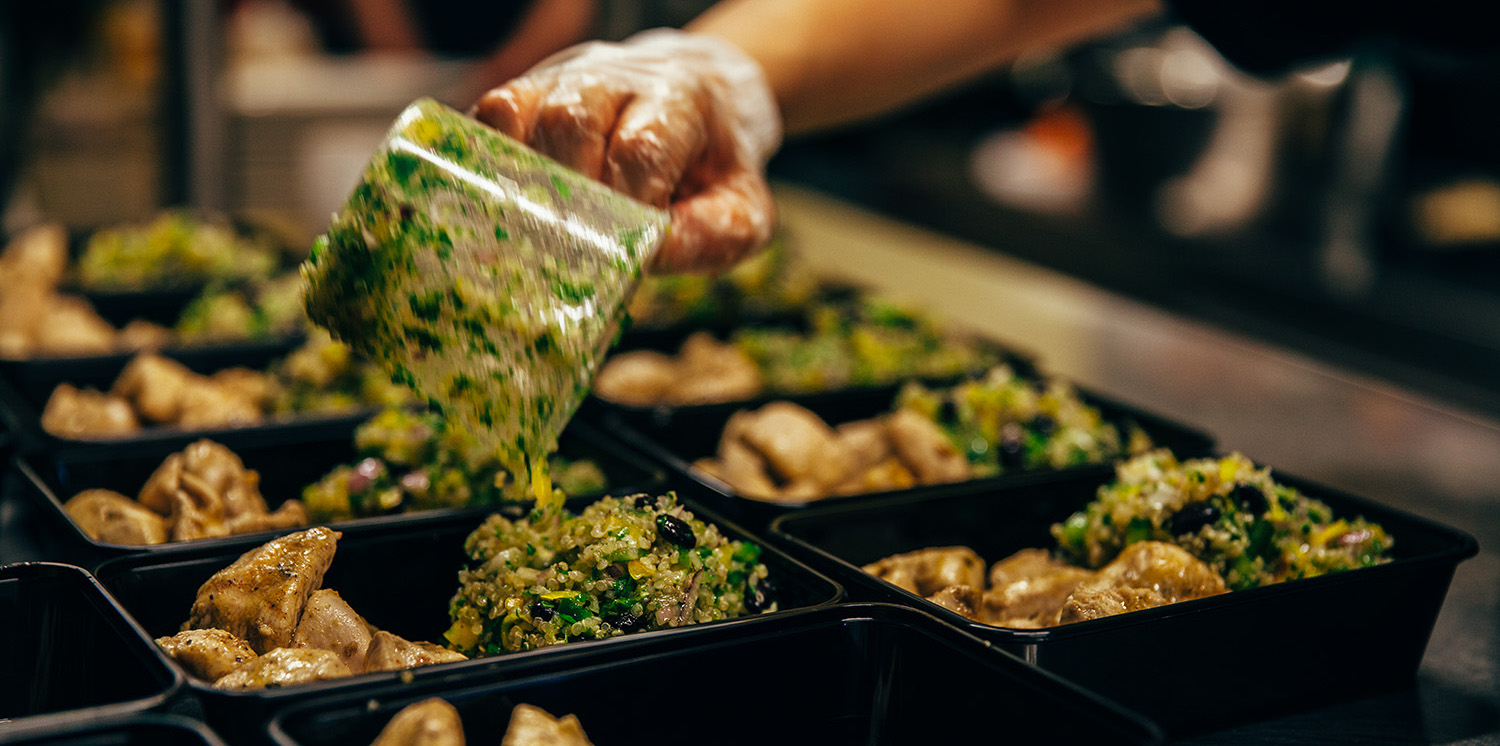 A photo of the chef in their professional kitchen, focused on the meals in their plastic black boxes, while the chef is pouring over a green herb sauce