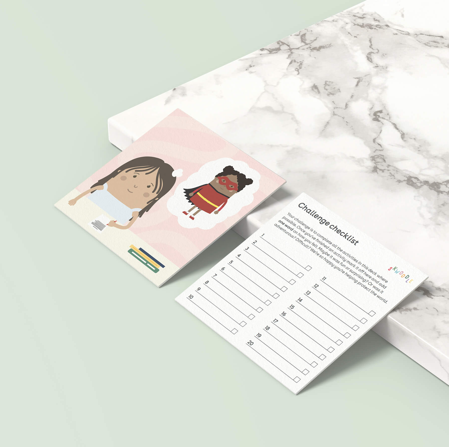 The front and back of one card propped up against a marble slab on a green slate background showing a girl in class dreaming of becoming a superhero