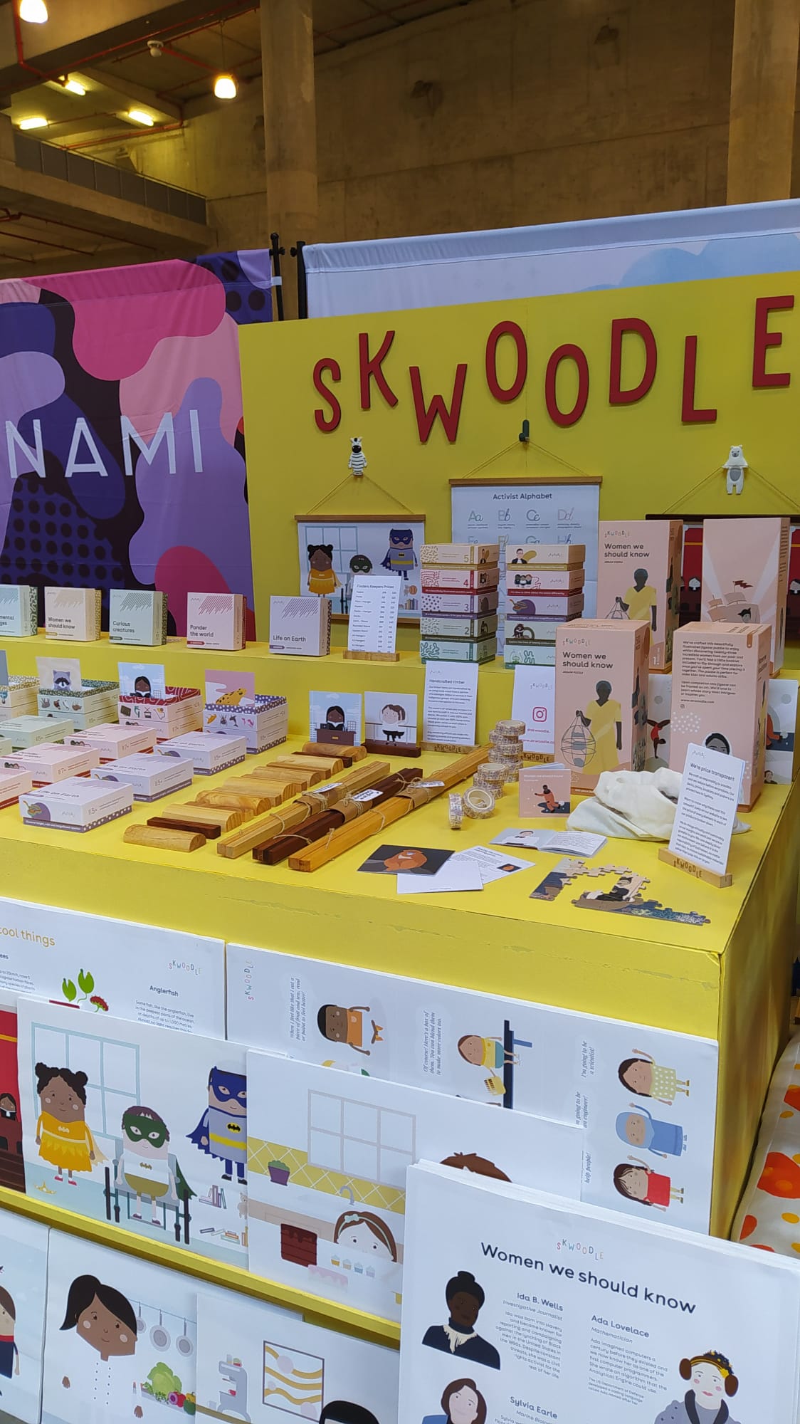 A bright yellow Skwoodle stall setup. The backboard hangs three posters and a 3D woodwork cut out of the SKWOODLE logo. On the table are the products, with a shelf in front for the posters and a heigher section on the table for the decks.