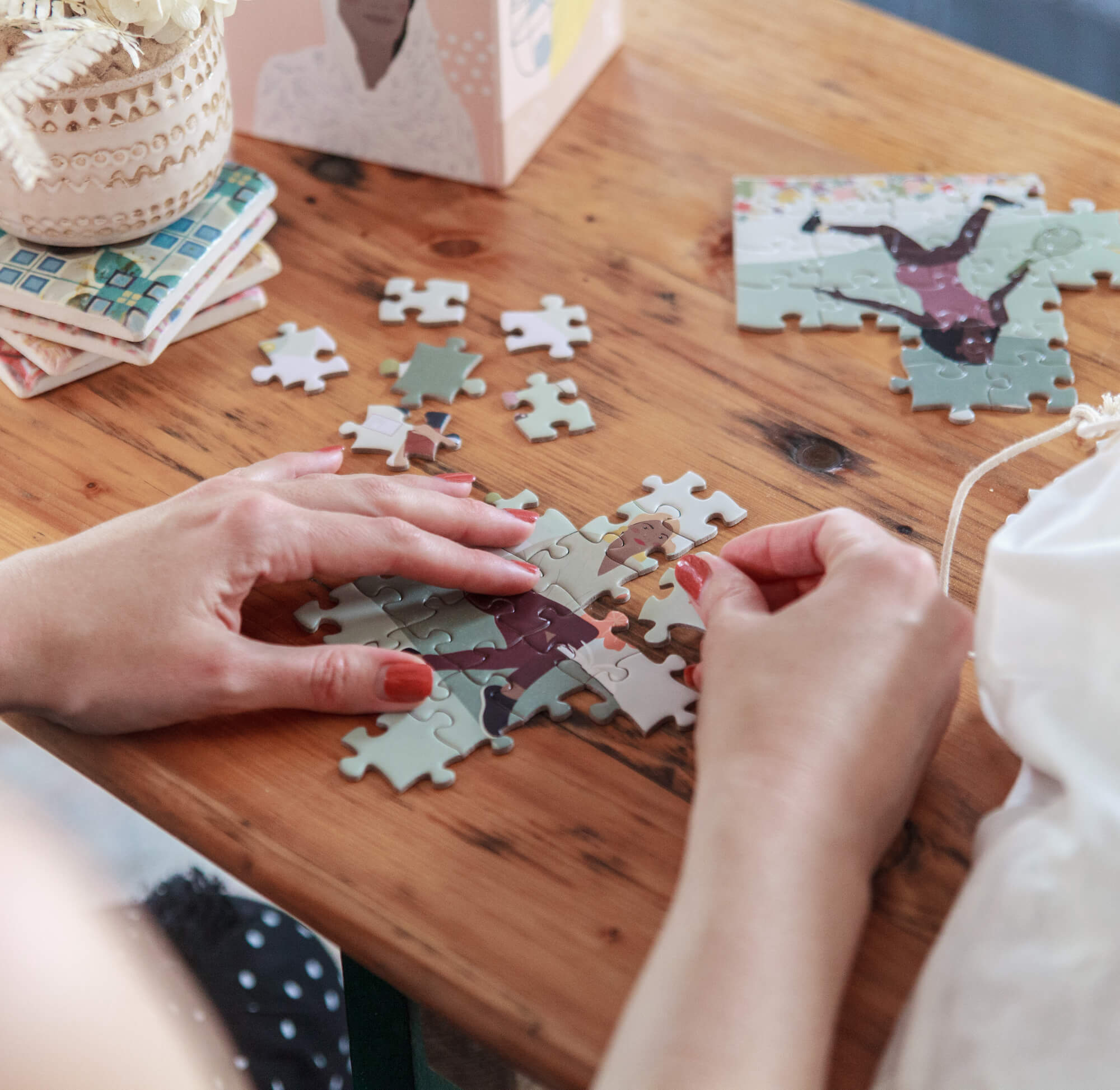 A photo of a woman making the puzzle. You can see her hands with red painted fingernails, laying puzzle pieces in their spots on a wooden table.