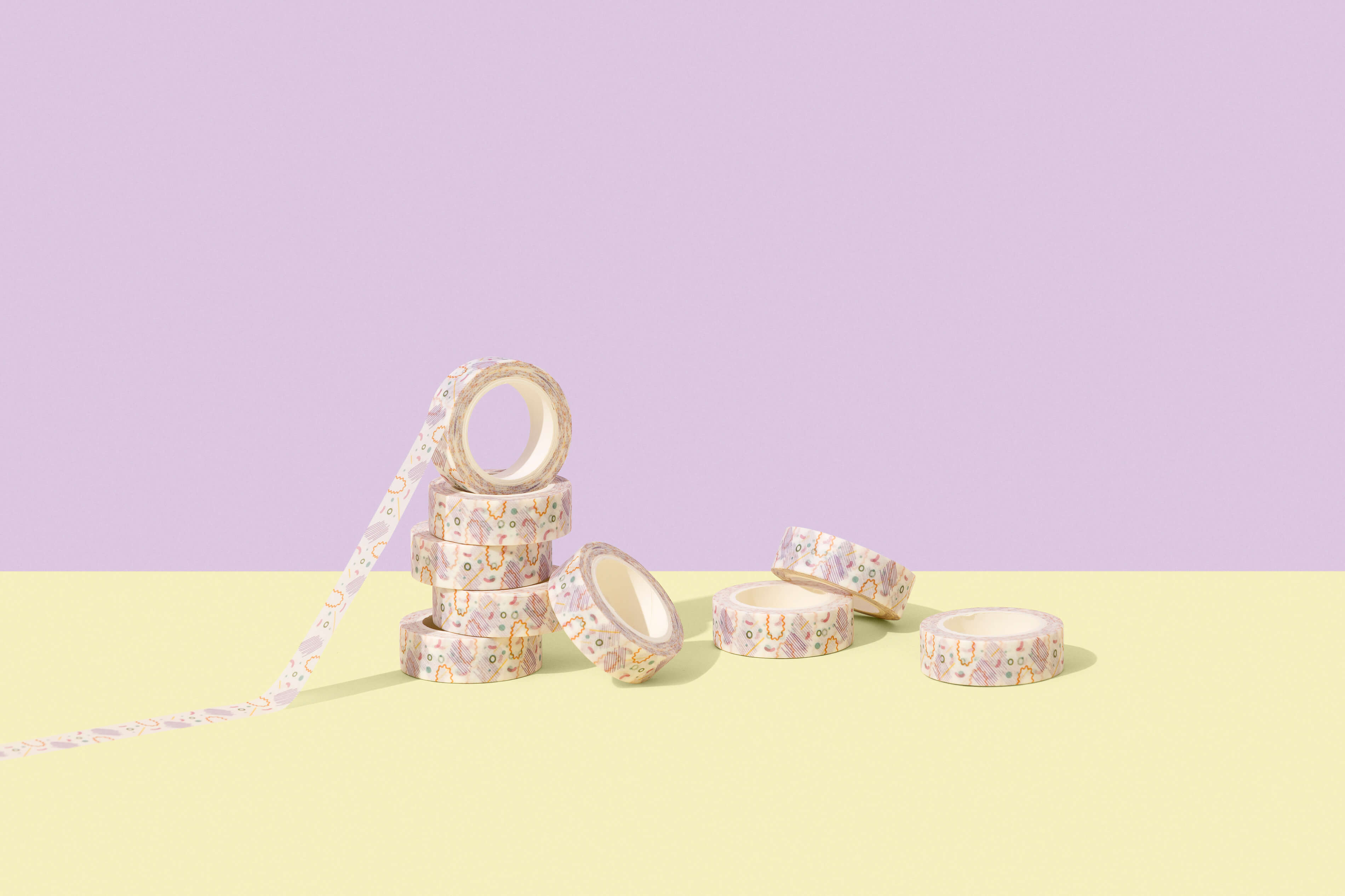 On a purple background with a yellow paper floor, is a stack of washi tapes with one of the top tapes pulled out and extended so you can see the Skwoodle pattern on it - this photo has been professionally shot and edited in a studio