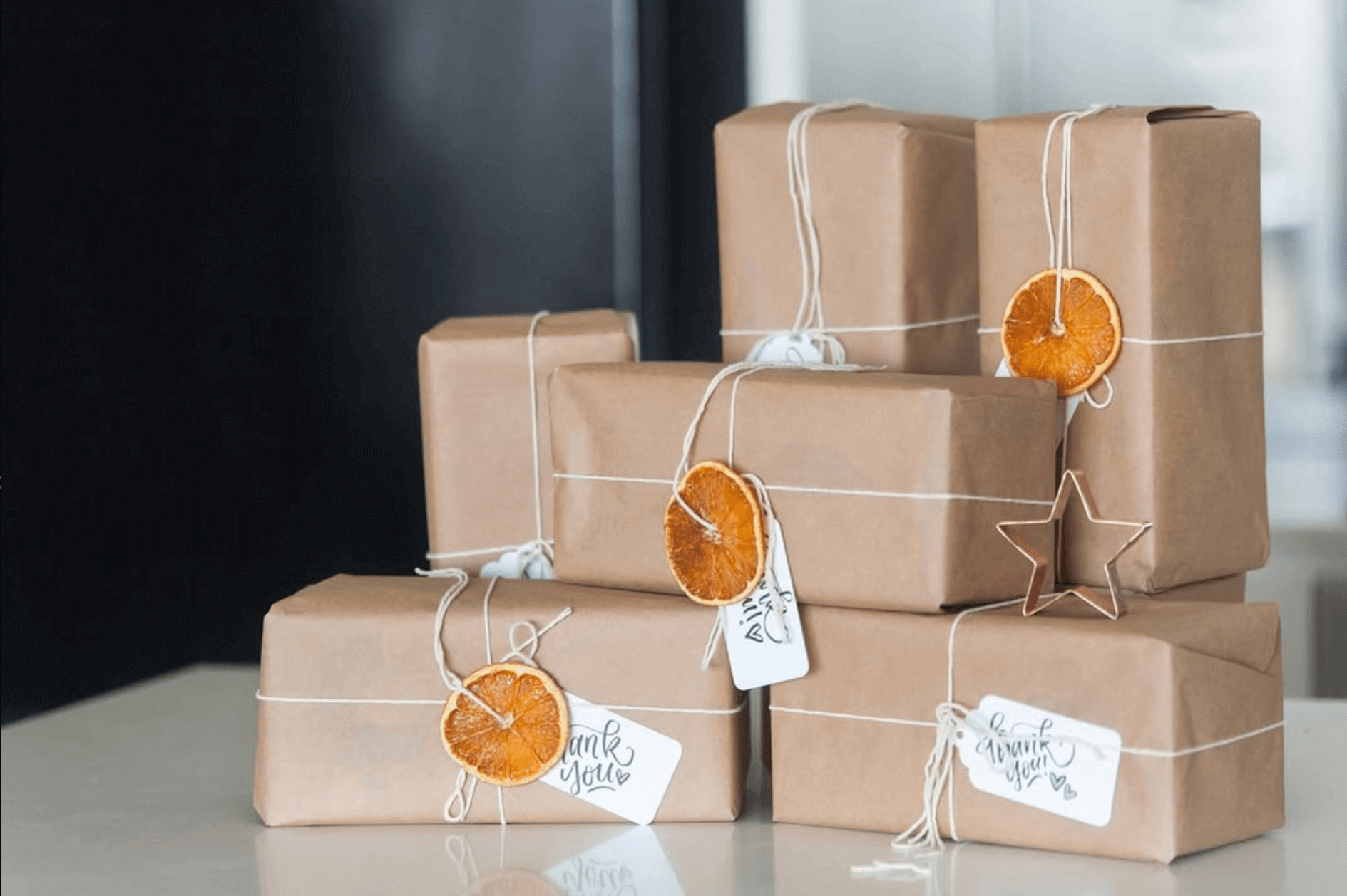 Puzzle boxes in brown paper are shown in a stack, each with twine around them, and a bow holding a handlettered thank you card and a dried orange for some festive holiday cheer